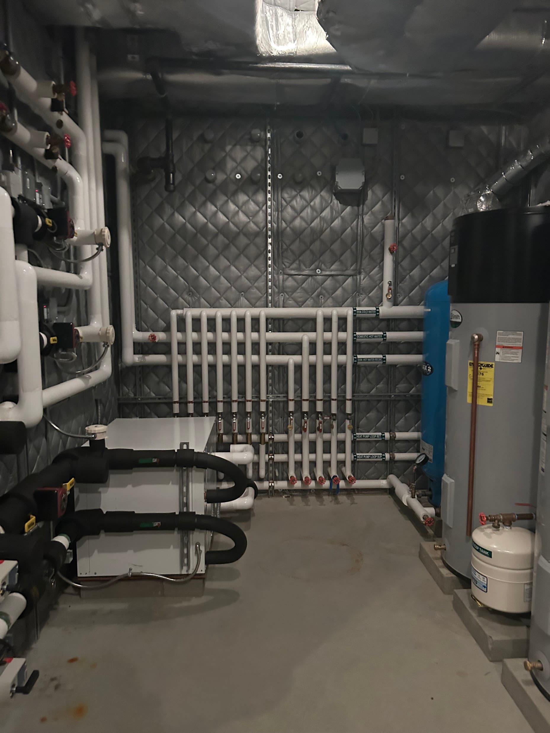 A fully finished mechanical room with a heat pump water heater, buffer tanks, and clearly labeled piping